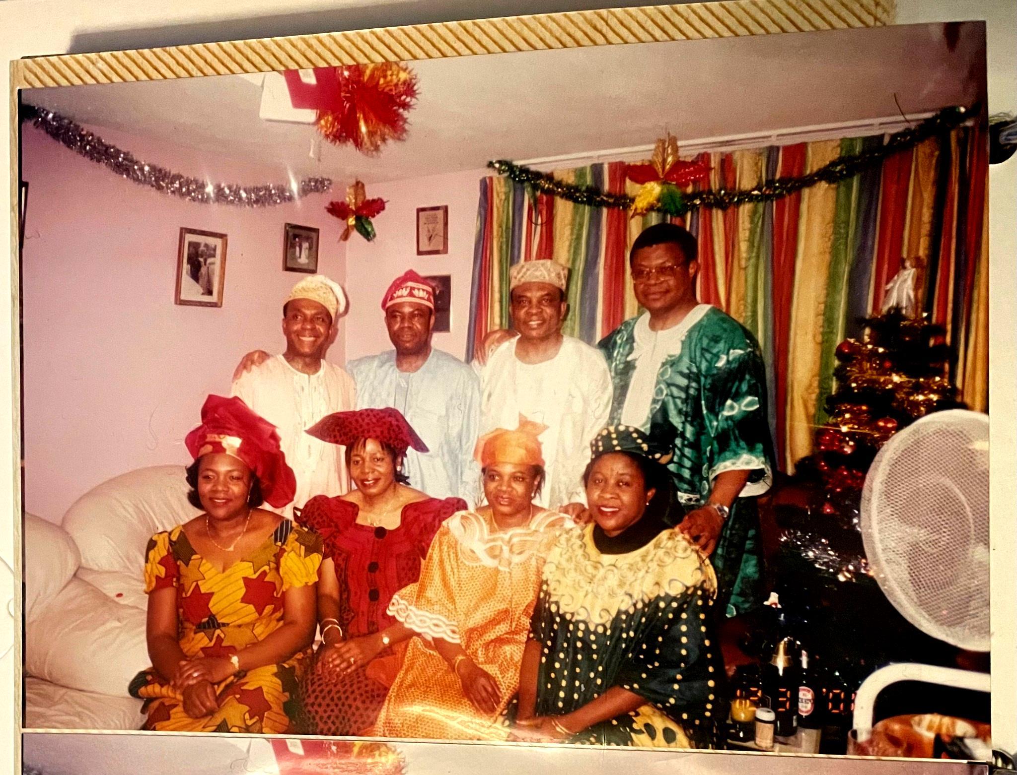 Uncles and aunties gathered at a New Year’s Eve party in London, smiling and dressed in colourful Nigerian clothing. Christmas decorations hang in the background, with drinks arranged in the foreground.